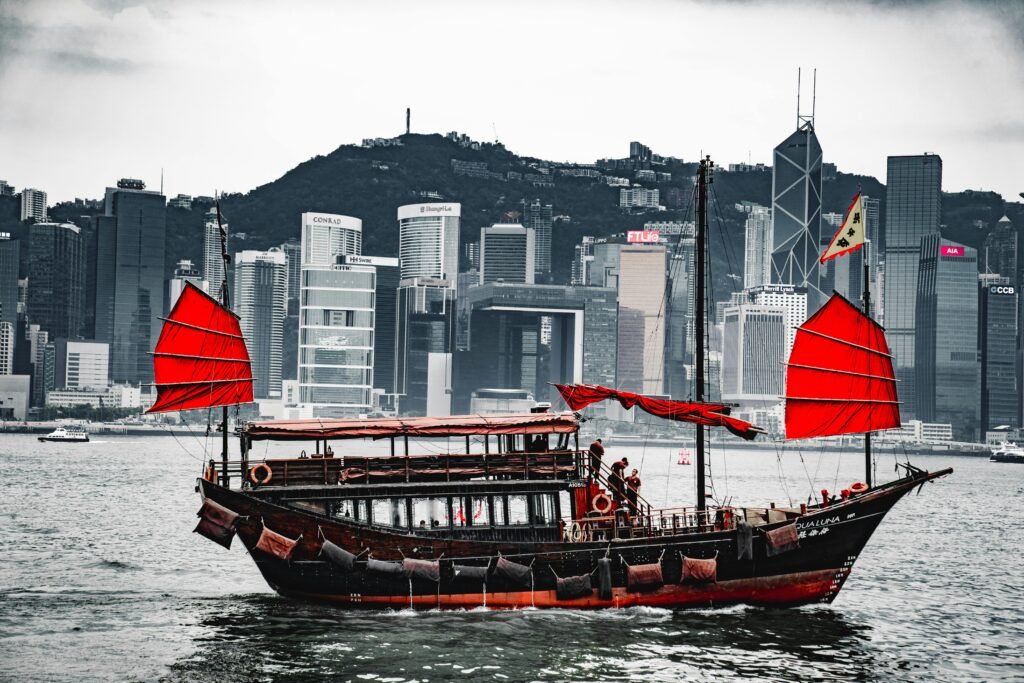 A traditional junk boat with red sails in Hong Kong harbor, against a skyline backdrop.